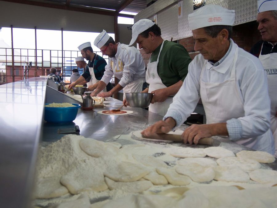 Volontari col cappello da cuoco stendono l’impasto della pizza su un lungo bancone infarinato